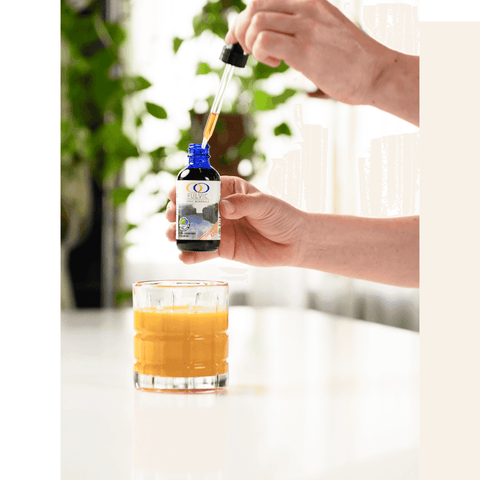 Hand holding Fulvic Ionic Minerals X350 bottle with a dropper over a glass of orange juice, demonstrating versatility in beverage mixing.