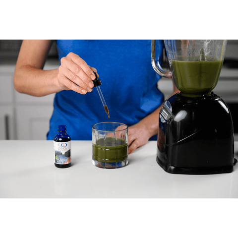 Person adding Fulvic Ionic Minerals X350 drops to a green smoothie, showcasing its compatibility with various health drinks.